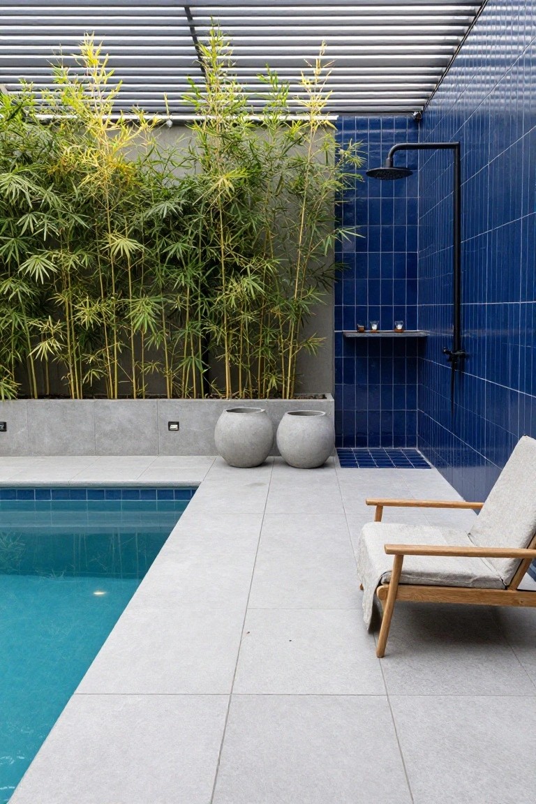 Above-ground pool adjacent to a blue-tiled outdoor shower with black fixtures, screened by bamboo plants, large gray pots, and a wooden lounge chair on a light gray tiled patio under a slatted roof.