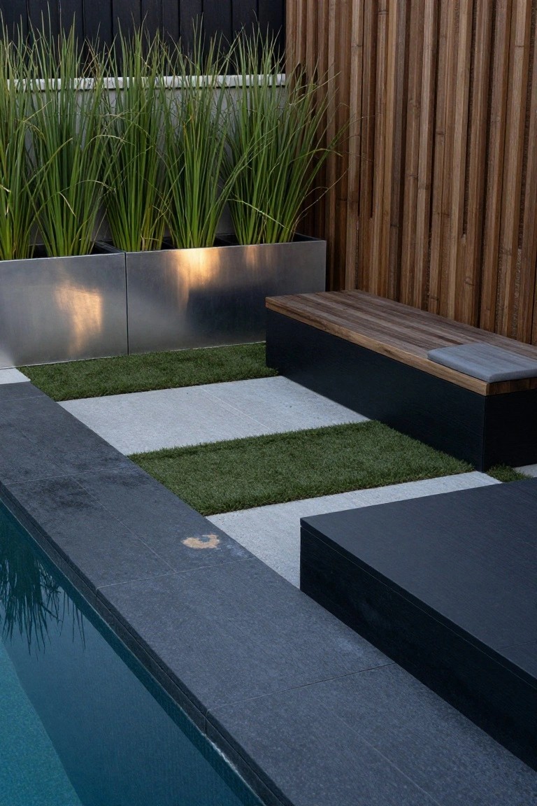 Close-up view of an above-ground pool edge with black tiled coping, a checkered pattern of green artificial grass and light gray concrete pavers, a black bench with walnut wood seat and gray cushion, stainless steel planters with tall grasses, and a wooden slat fence in the background.
