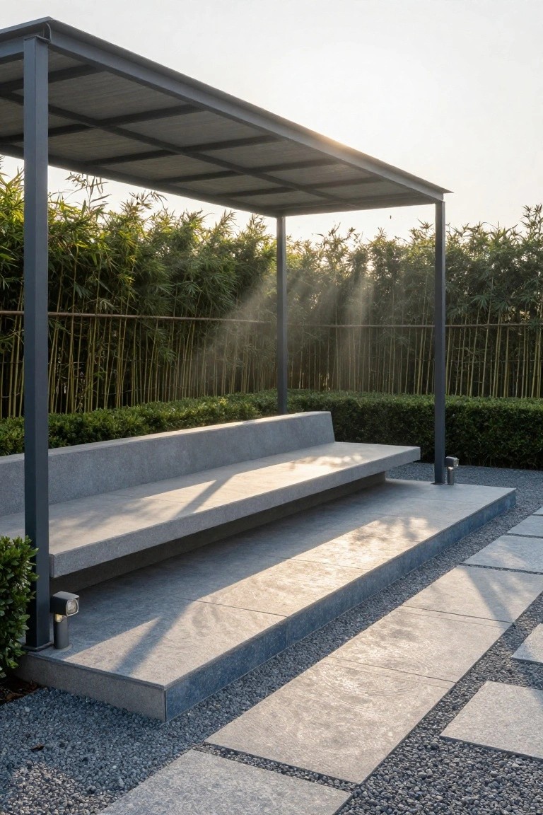 Metal-roofed open shelter covering a long gray stone bench with steps, backed by tall bamboo screen and flanked by low green hedges on a gravel and stone paver terrace.