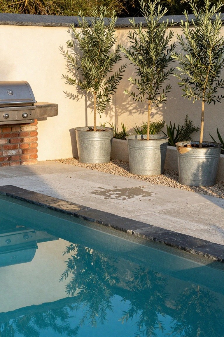Poolside stone paving with gravel mulch, a built-in brick barbecue, and three large galvanized metal buckets planted with olive trees next to a turquoise pool.
