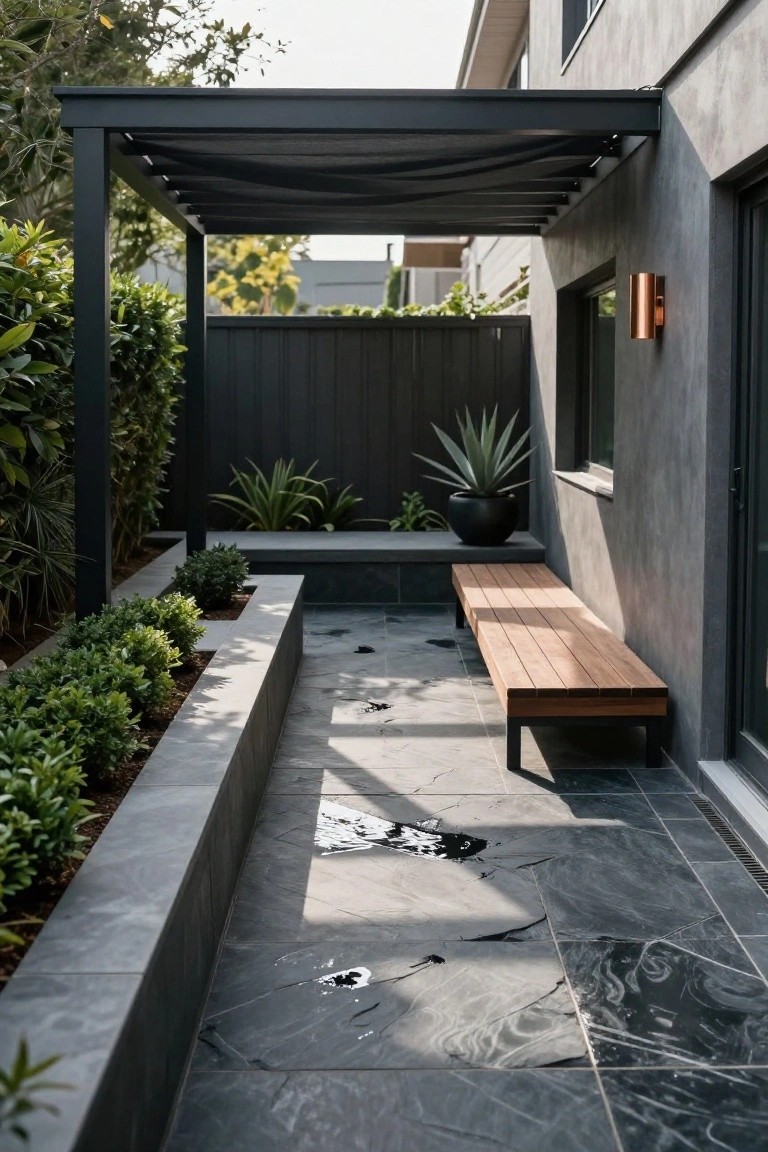 Modern outdoor patio with long wooden bench on dark slate tiles, stone wall planters filled with green shrubs, large black potted agave plant, black metal pergola with shade cloth, and house wall with copper light fixture.