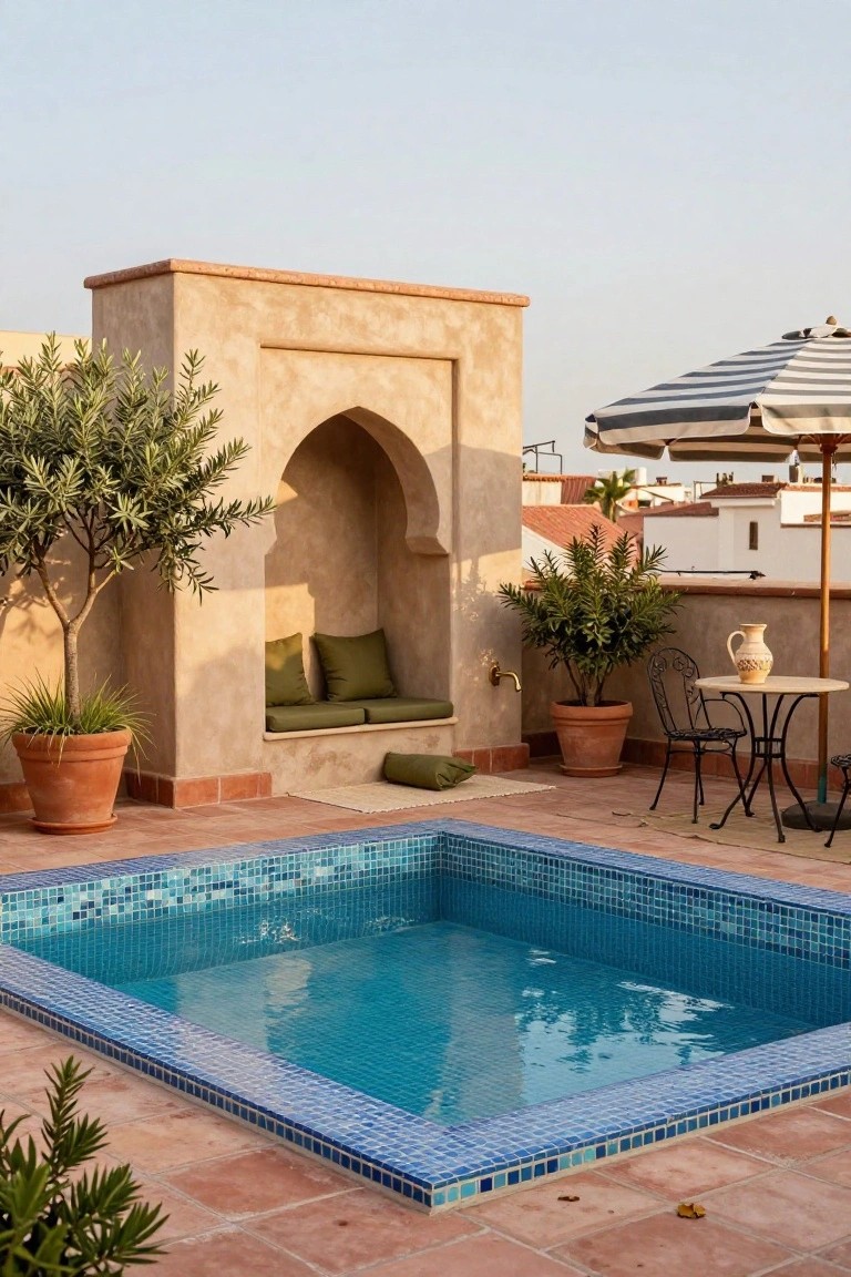 15 Rooftop Pool Design Ideas for Sky-High Luxury Rooftop terrace with small blue mosaic plunge pool edged in terracotta tiles, arched beige wall niche containing green cushioned bench and pillows, potted plants including olive trees, small round table with wrought-iron chairs under striped umbrella.