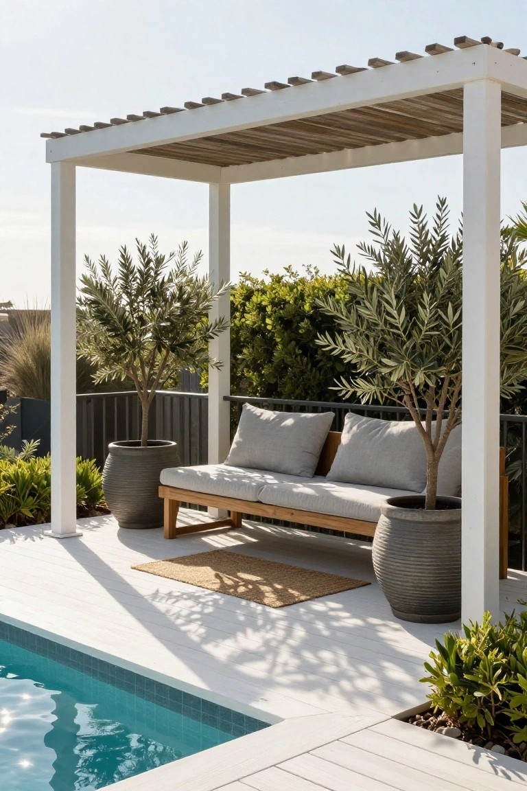 White pergola with wooden slats covers a wooden bench with gray cushions on a light deck next to a blue pool, flanked by large potted olive trees in gray pots.