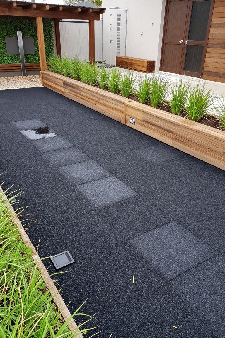 Outdoor terrace with black rubber square tiles as the main deck surface, a linear path of gray pavers, wooden raised planters with tall grasses on both sides, a pergola overhead, wooden bench, and nearby white building wall with wood-clad door.