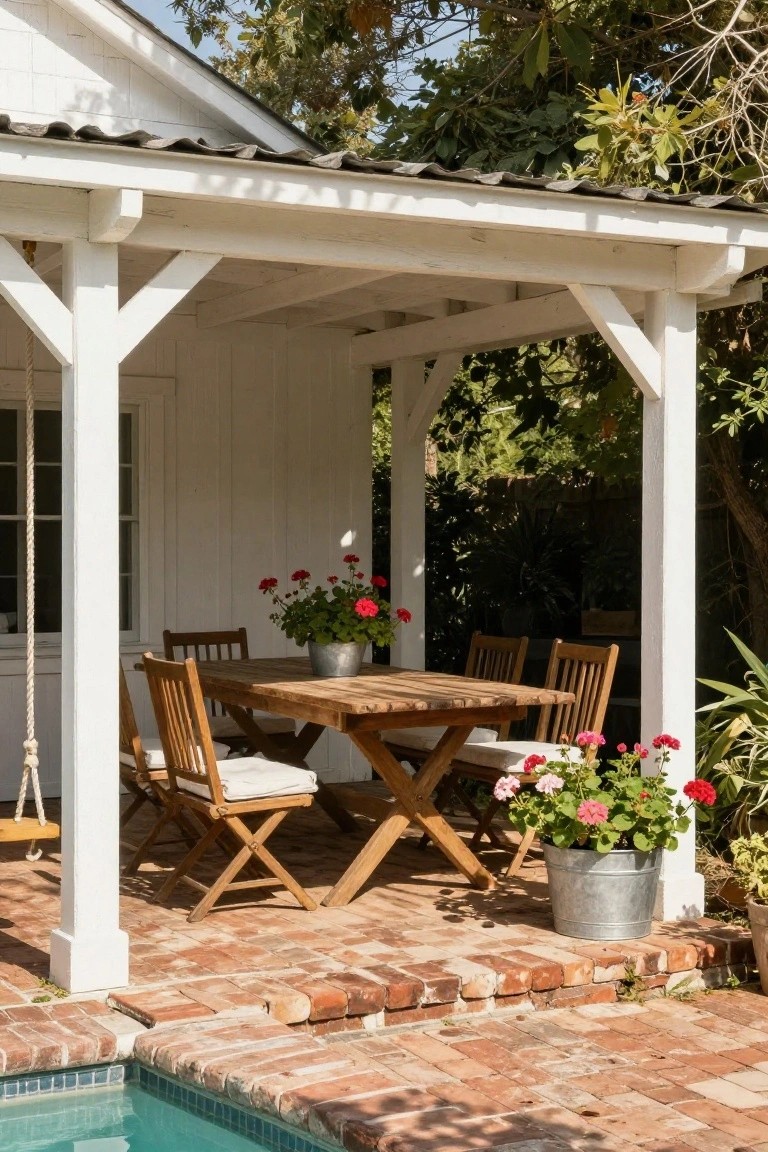 17 Elegant Pool Deck Pergola Designs That Add Structure and Shade White wooden pergola with shingle roof covering a rectangular wooden dining table and four chairs on brick pavers next to a turquoise pool, with red geraniums in metal buckets on and around the table.