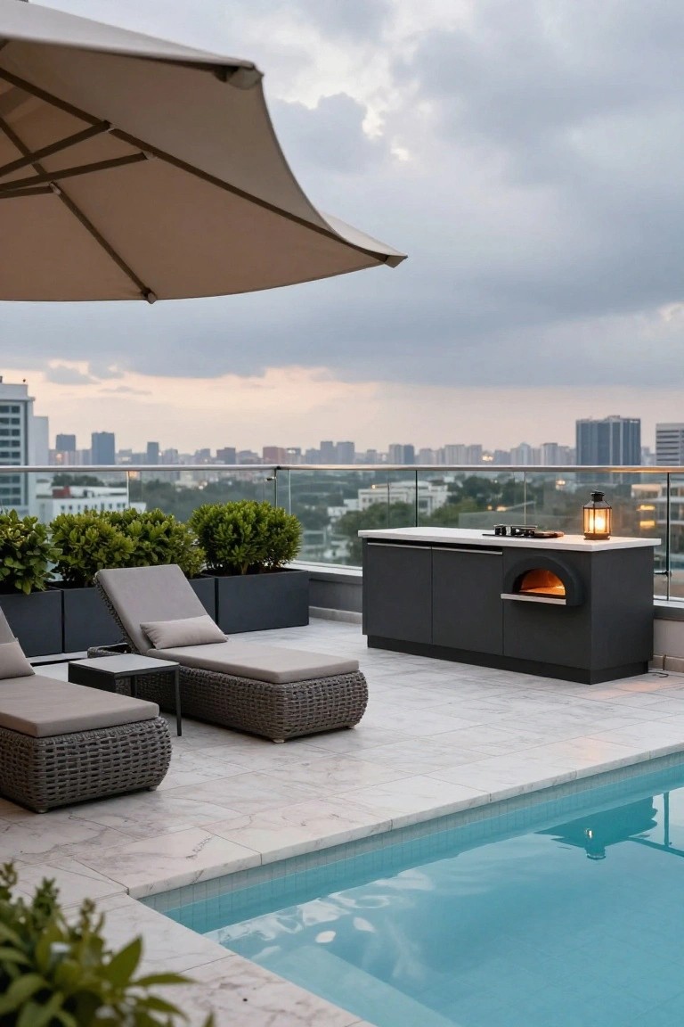 Rooftop terrace with turquoise inground pool, two beige lounge chairs on white marble tile deck, black outdoor kitchen cabinetry featuring wood-fired pizza oven, potted green shrubs, beige umbrella, glass railing, and city skyline under cloudy sky.