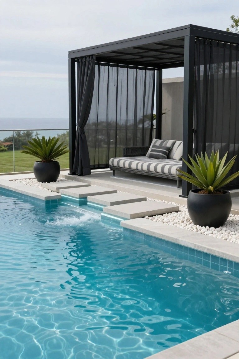 17 Modern Inground Pool Deck Ideas That Define Luxury Black metal pergola with sheer curtains and a striped daybed positioned beside an inground pool with stone steps, white pebbles, black pots of agave plants, and an ocean view in the background.