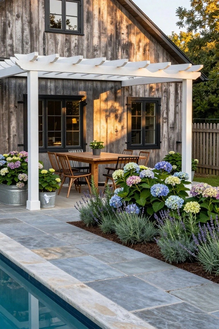 Weathered dark wood barn with attached white pergola covering wooden dining table and chairs on stone patio, potted hydrangeas nearby, adjacent to rectangular in-ground pool with gray stone decking.
