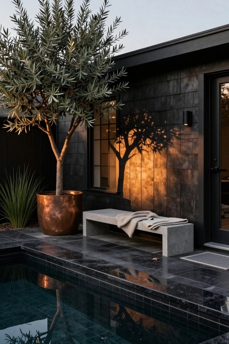 Poolside concrete bench with beige throw blanket next to a potted olive tree in copper container against black shingled wall, with dark tiled pool edge in foreground.