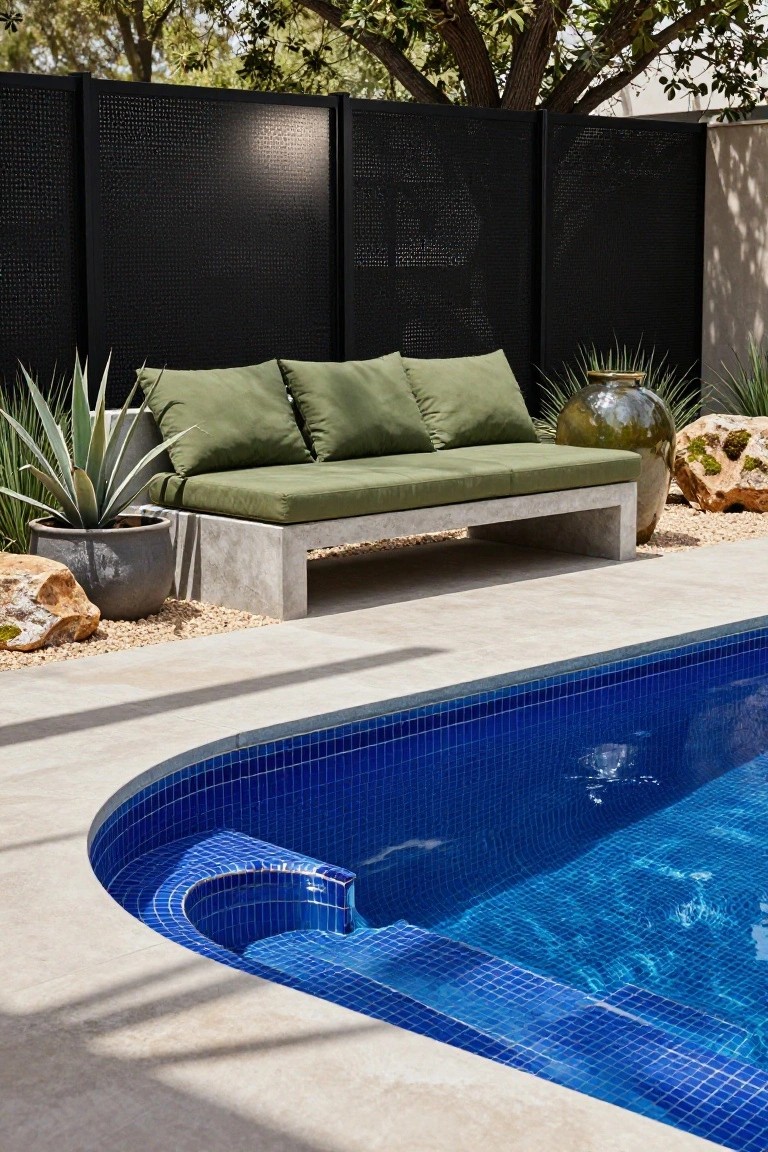 A low concrete bench with thick green cushions sits poolside next to potted agave plants, large pottery, rocks, and gravel, adjacent to a curved blue mosaic-tiled pool edge with black privacy screens and trees behind.
