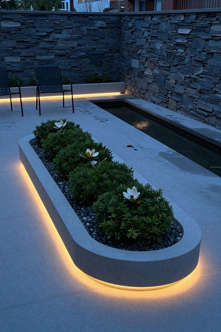Oval concrete above-ground planter filled with green shrubs, white flowers, and black pebbles, with yellow LED lighting along the lower edge, on a gray paved patio beside a narrow water channel and flanked by stone walls and two chairs.