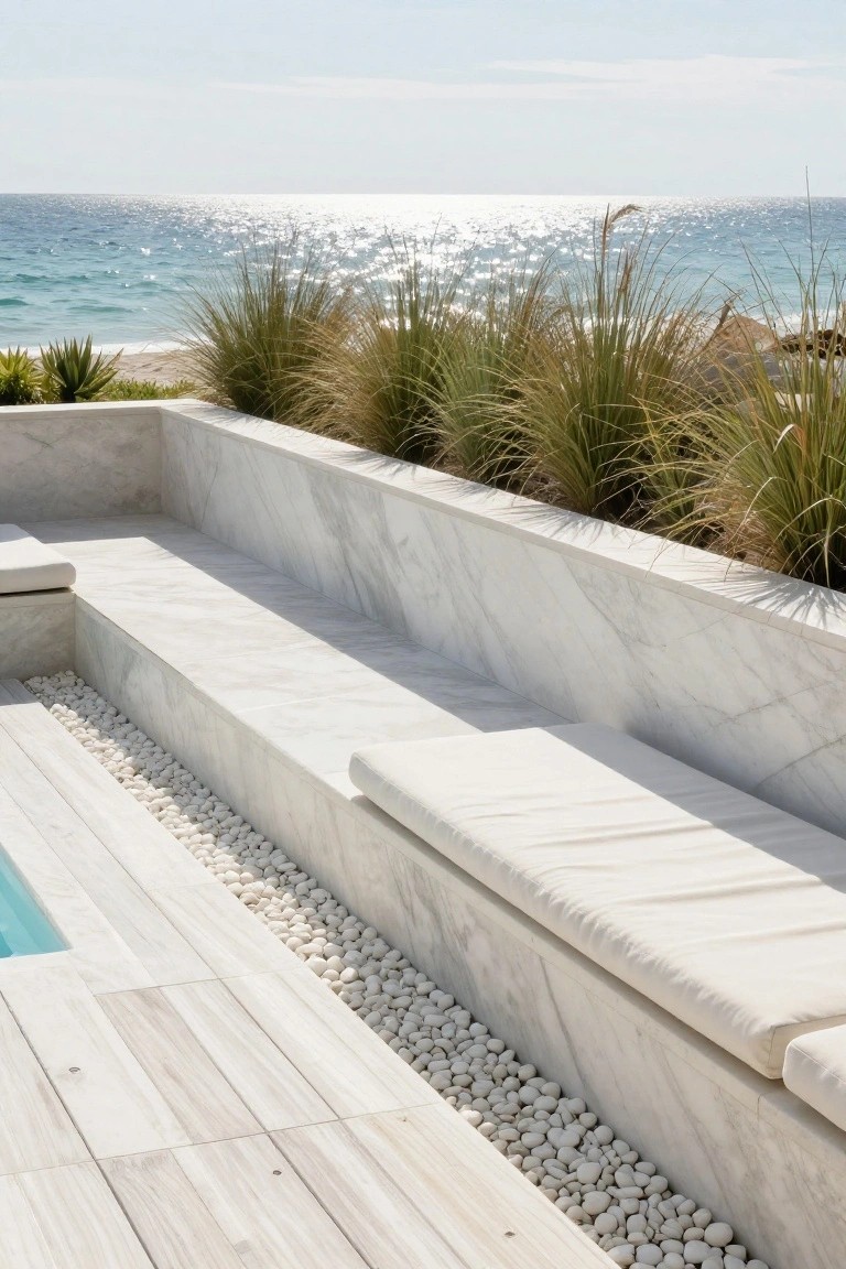 Pool deck with light wood-grain porcelain tiles, white marble built-in benches with cushions, pebble-filled edging strip, and tall grasses near the ocean.