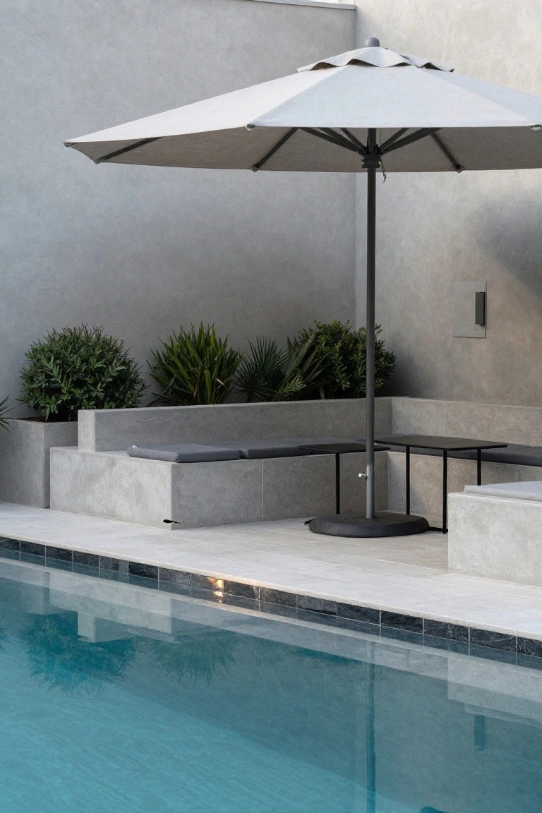 A beige cantilever umbrella shades low gray concrete benches and small black tables next to a rectangular pool with black tile edging, potted plants along a rendered wall.