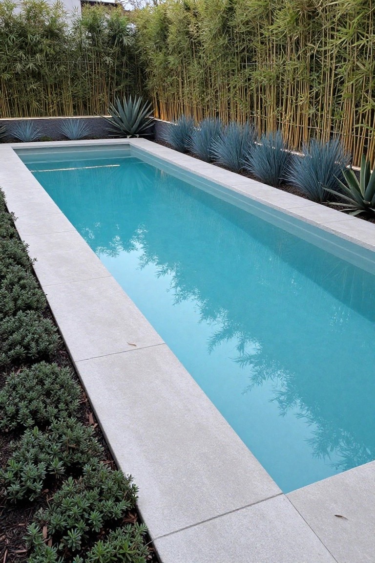 Narrow rectangular pool with turquoise water and white stone coping, bordered by tall bamboo screening on one side, blue agave plants along the edges, and low green shrubs on the other side.