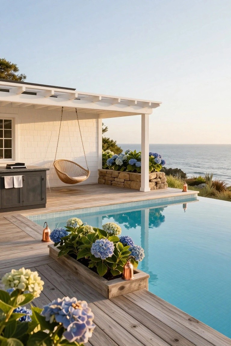 Wooden deck around turquoise infinity pool with raised planters of blue and white hydrangeas, woven hanging swing under white pergola, dark cabinetry with grill, stone retaining wall, and ocean view in background.
