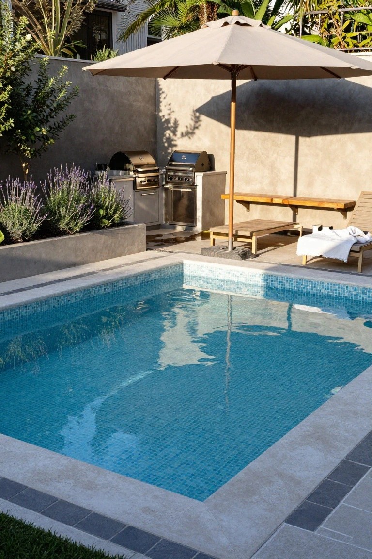 Small rectangular inground pool with turquoise mosaic tiles and travertine coping, adjacent to built-in stainless steel BBQ grills on a raised concrete platform, with wooden loungers, beige umbrella, lavender planters, and textured walls in a sunny backyard.