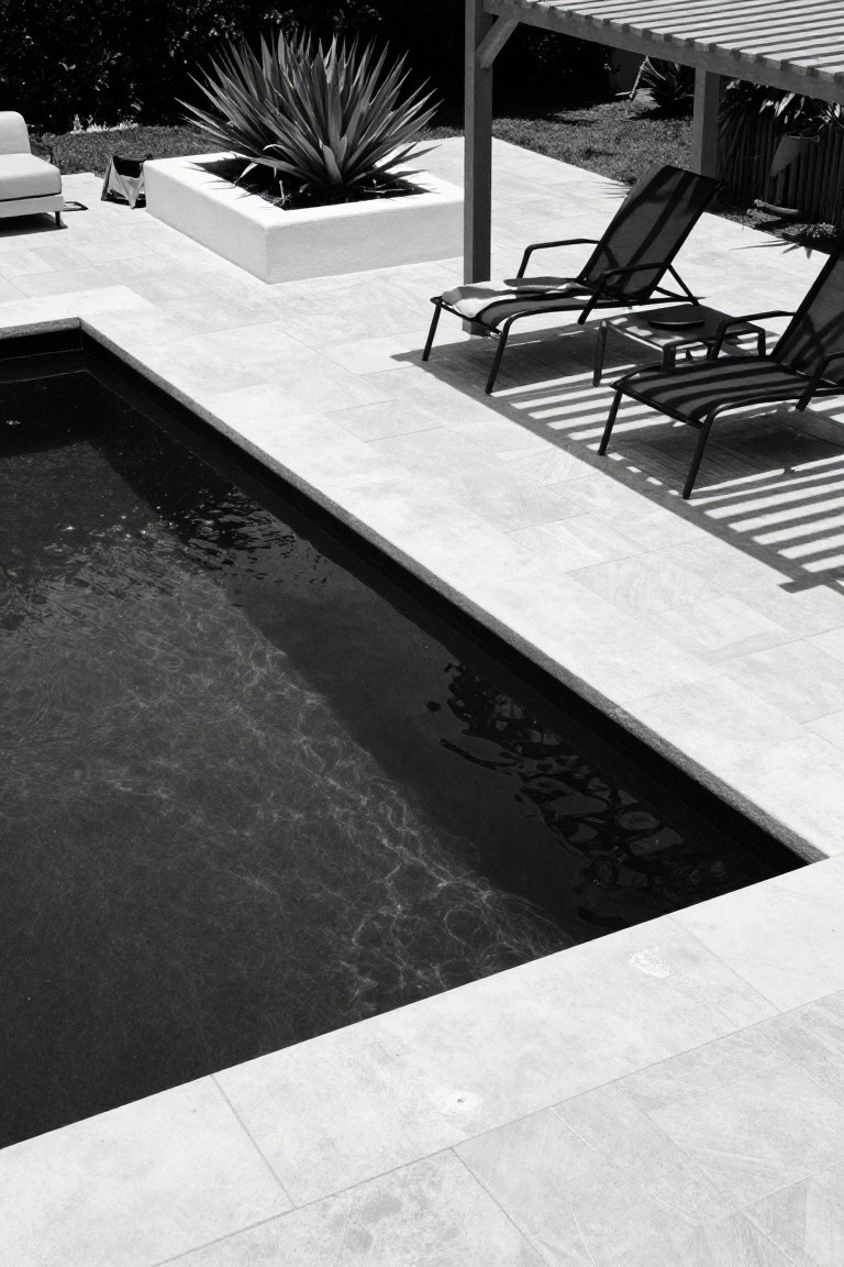 Black-and-white photo showing a small rectangular inground pool with dark rippling water, edged by light gray-white stone decking, two lounge chairs under a wooden pergola, a potted agave plant, and nearby outdoor sofa.