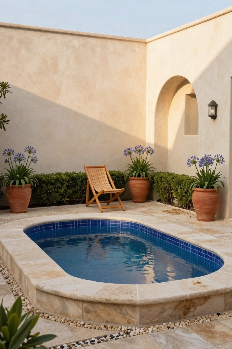 Small oval inground pool with blue mosaic tiles in a beige stucco courtyard surrounded by terracotta pots of agapanthus, boxwood shrubs, a wooden folding chair, and pebble accents.