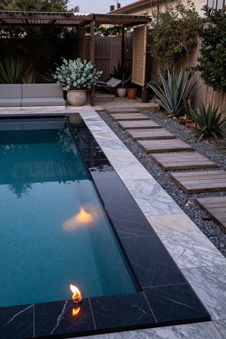 Small rectangular inground pool with black marble coping holding a flame burner on one edge, white marble tiles nearby, wooden stepping stones on gravel, succulents, lounge seating, and pergola in a backyard at dusk.