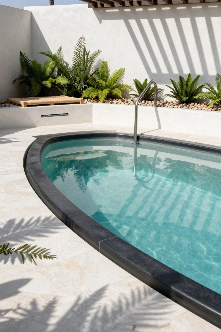 Oval turquoise pool with black edging on white stone decking surrounded by ferns, pebbles, and a wooden bench in a minimalist outdoor setting.