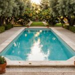 Turquoise pool with travertine edging and pebble borders, flanked by potted rosemary plants, a rustic wooden bench, terracotta pots, and overhanging olive trees with fruit amid lemon trees and garden hedges.