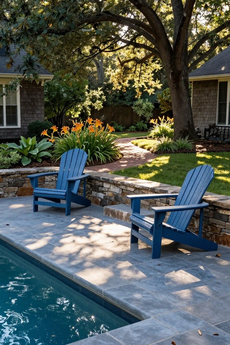 Two blue wooden Adirondack chairs positioned on a gray slate tile pool deck next to a rectangular swimming pool, backed by a low stone retaining wall, with landscaping and trees nearby.