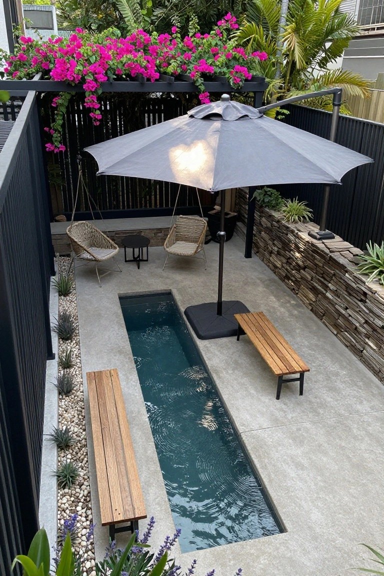 Overhead view of a narrow rectangular plunge pool in a concrete-paved courtyard, flanked by wooden benches, rattan hanging chairs, a gray cantilever umbrella, black fences, stone walls, and tropical plants.
