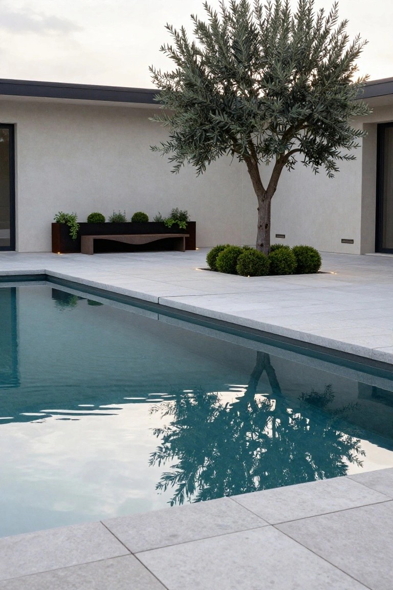 Modern infinity-edge pool with turquoise water and white stone decking, olive tree at the edge surrounded by boxwood shrubs, curved corten bench against white stucco wall, and soft uplighting.