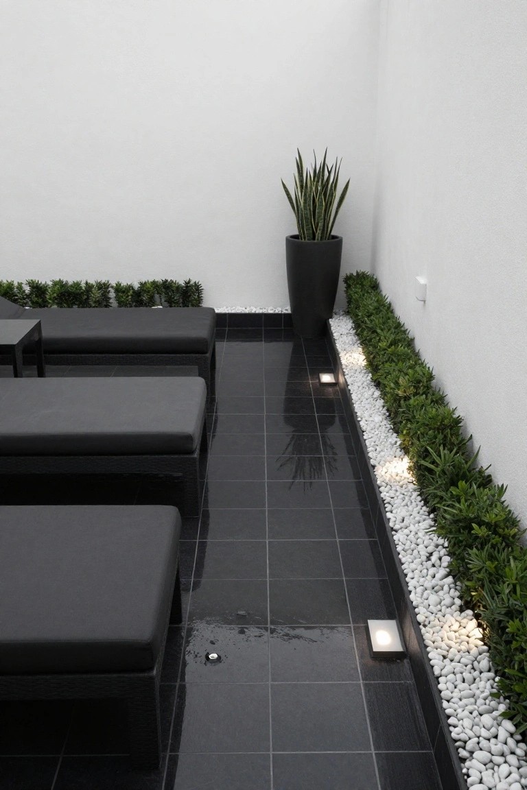 Outdoor terrace with black sun loungers on glossy black tiles, bordered by a strip of white pebbles and low green boxwood hedges against a white wall, featuring recessed lights and a black potted snake plant.