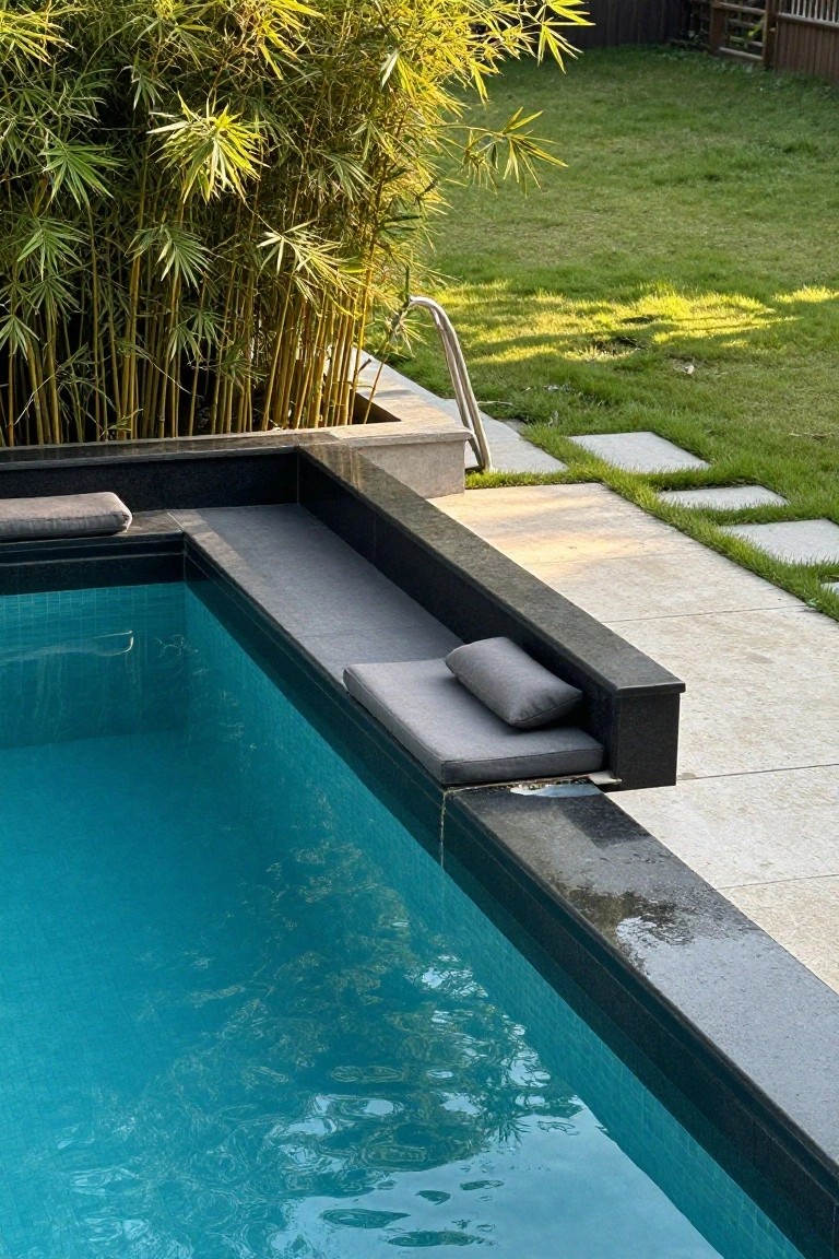 Close-up of a pool edge with built-in black stone bench topped by gray cushions, bamboo plants to one side, pool ladder, and adjacent paver path and grass.
