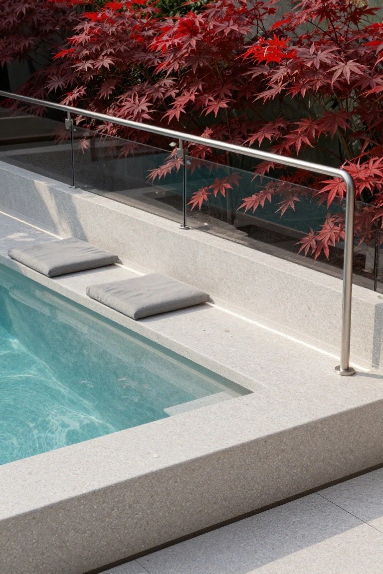 Modern pool with white terrazzo decking, two gray cushions on built-in edge seats, stainless steel and glass railing, and red maple shrubs in the background.