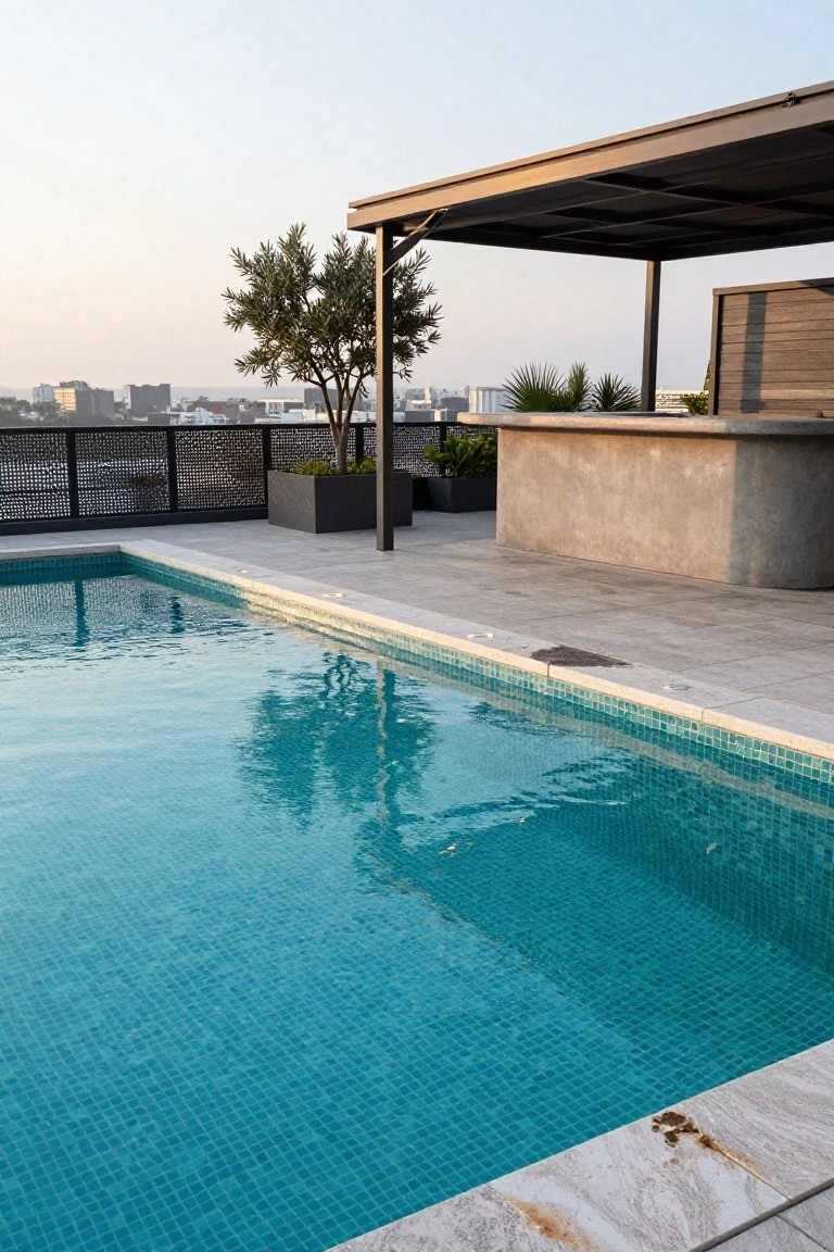 Rooftop pool with turquoise mosaic tiles, light stone decking, concrete bar counter, potted plants, metal pergola, and distant city view.