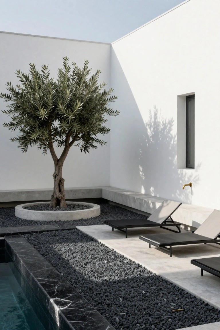 18 Sophisticated Landscaping Around Inground Pool Designs That Flow Beautifully White minimalist building corner featuring an olive tree in a circular concrete planter surrounded by black gravel, with lounge chairs nearby and a black marble-edged inground pool.
