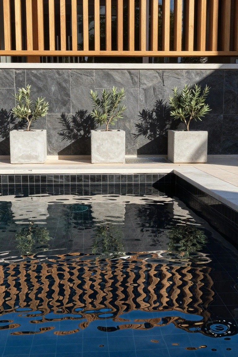 Black mosaic-tiled swimming pool with reflections of wooden balustrade, potted olive trees, and dark stone wall, edged by beige tiles.
