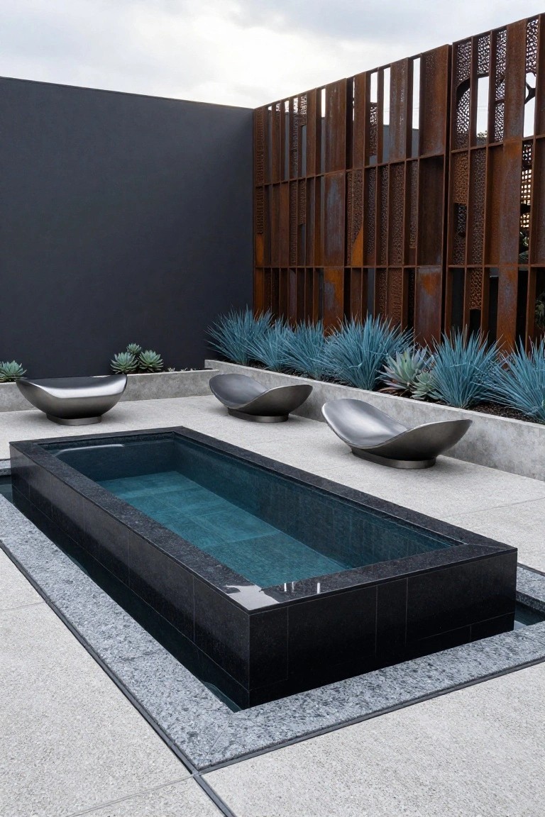 Narrow rectangular inground pool with black tile edges on a concrete patio, surrounded by three metallic bowl-shaped chairs, blue agave plants, and a rusted metal screen fence against a dark wall.