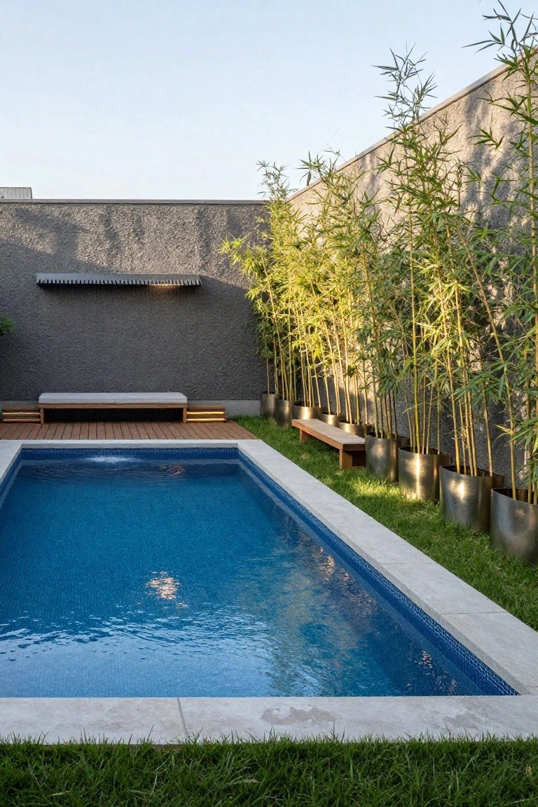 Rectangular inground pool with blue mosaic tiles and white coping, bordered by grass and a wooden deck with a cushioned bench, with tall bamboo plants in large shiny metal pots lined up along one edge against a textured gray wall.