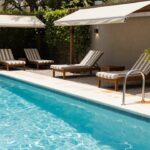 A turquoise pool with a stainless steel ladder at the edge, wooden lounge chairs with striped cushions along the side, under a wooden pergola with beige fabric shades, surrounded by hedges and potted plants.