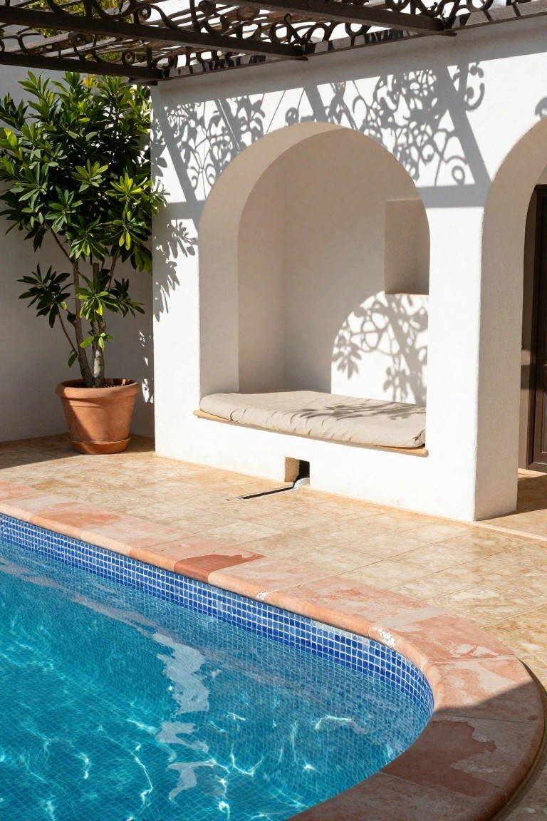 White stucco wall with arched niche containing a cushioned bench seat next to a curved blue mosaic pool edged in terracotta tiles on beige decking, potted plant nearby and wrought iron pergola overhead.