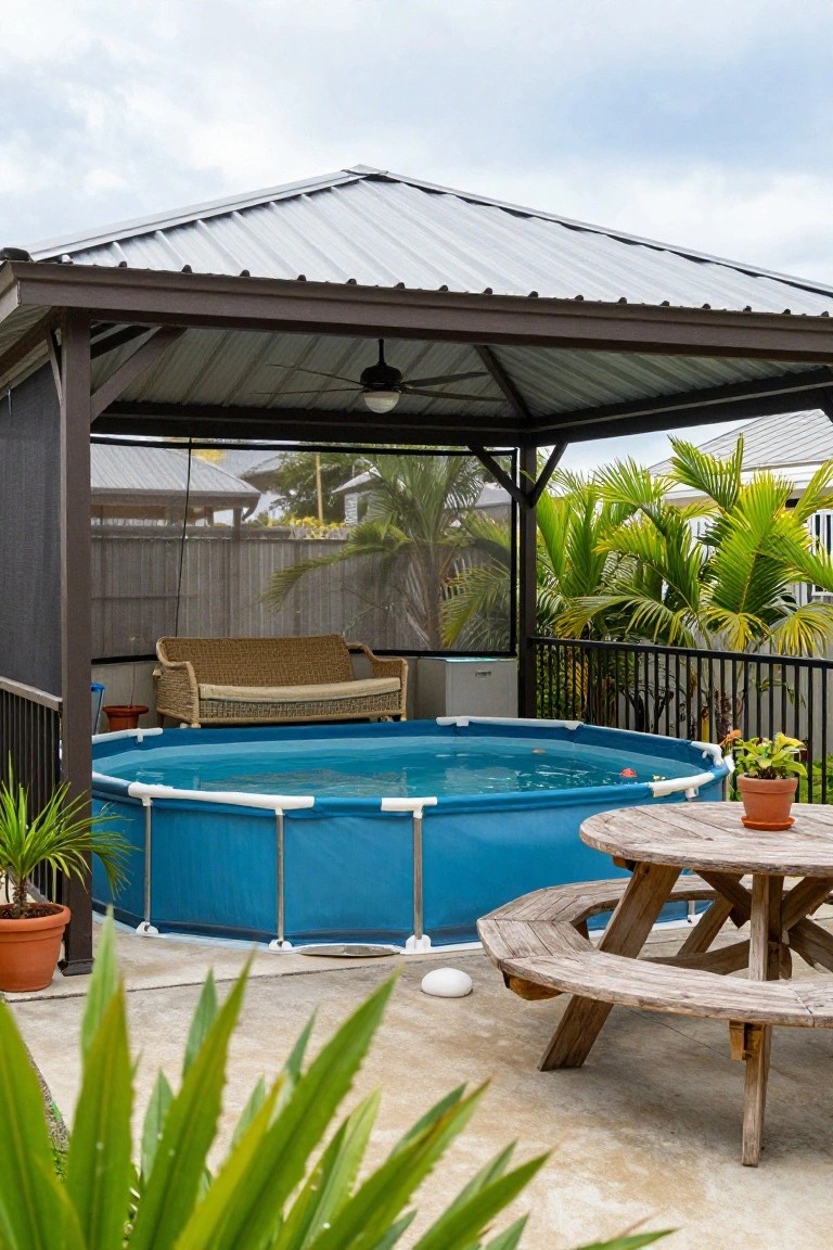 Above-ground blue pool inside a metal-roofed screened pergola with ceiling fan, wicker sofa, wooden picnic table, potted plants, and tropical landscaping nearby.