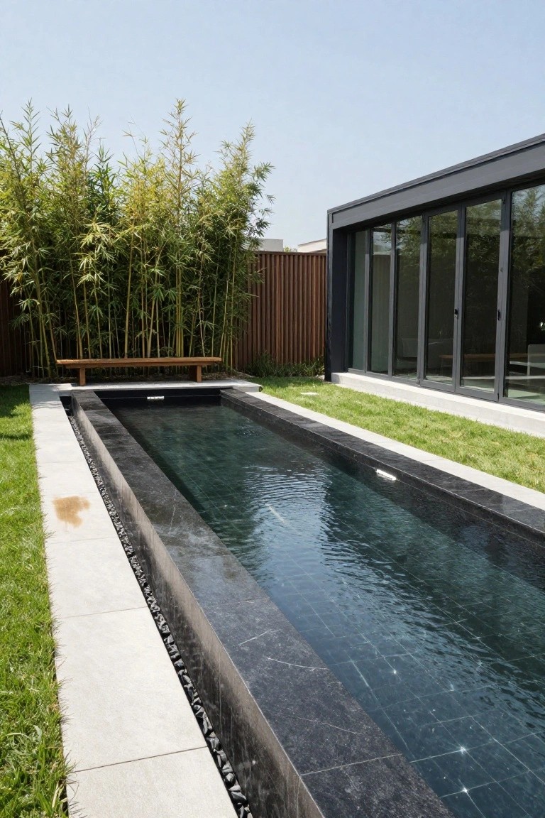 Narrow elongated inground pool with black stone edging in a grassy yard, wooden bench opposite bamboo screen, next to modern house with large sliding glass doors.