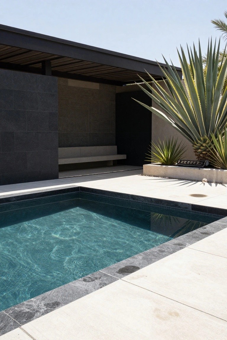 Square pool with dark gray stone edging and teal rippling water on a light stone deck, next to a built-in bench under a wooden pergola, raised planter with agave plants, and textured black walls.