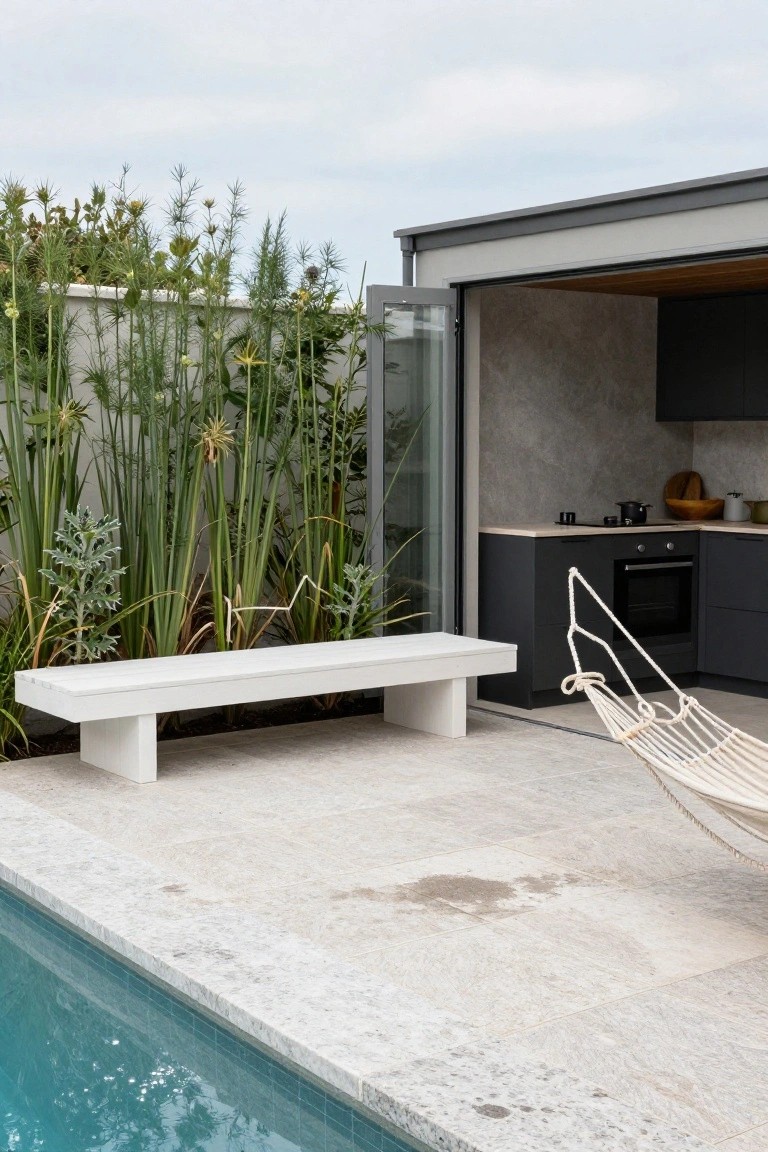 White rectangular bench on tiled patio next to turquoise pool edge, tall green reeds and plants against white wall, open folding glass doors revealing modern kitchen with black cabinets and hanging hammock.