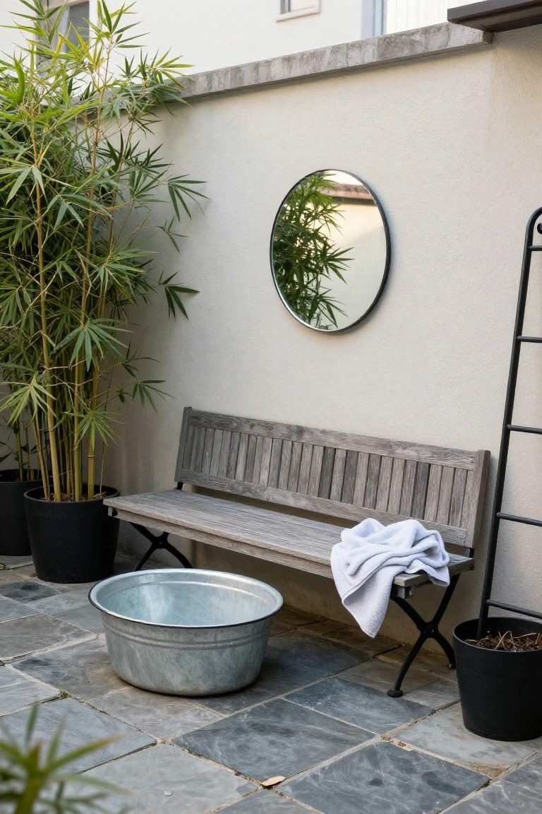 20 Compact Stock Tank Pool Ideas Perfect For Small Spaces Small outdoor courtyard with tall potted bamboo plants against a light wall, round mirror, wooden bench draped with white towel, black metal ladder shelf, large galvanized metal tub on gray stone pavers, and additional potted plants.