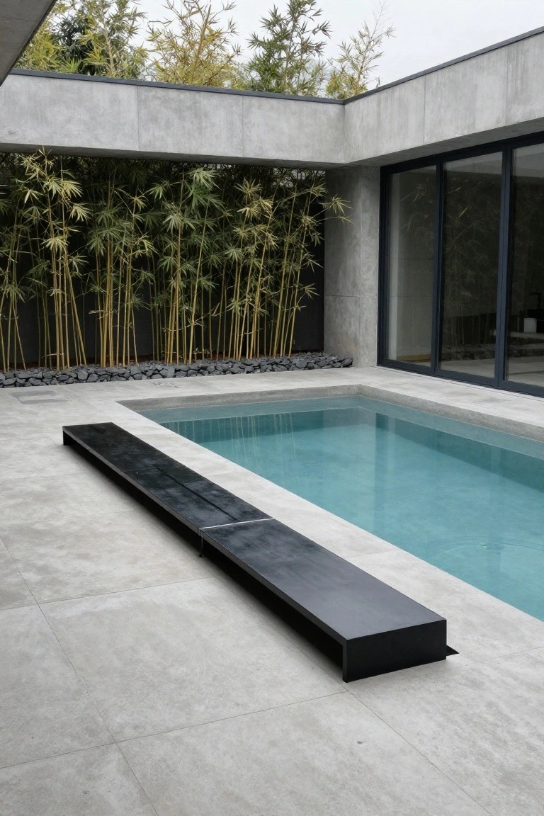 Narrow inground pool with turquoise water next to a long black rectangular bench on gray concrete deck, backed by bamboo plants and concrete walls with large glass doors.