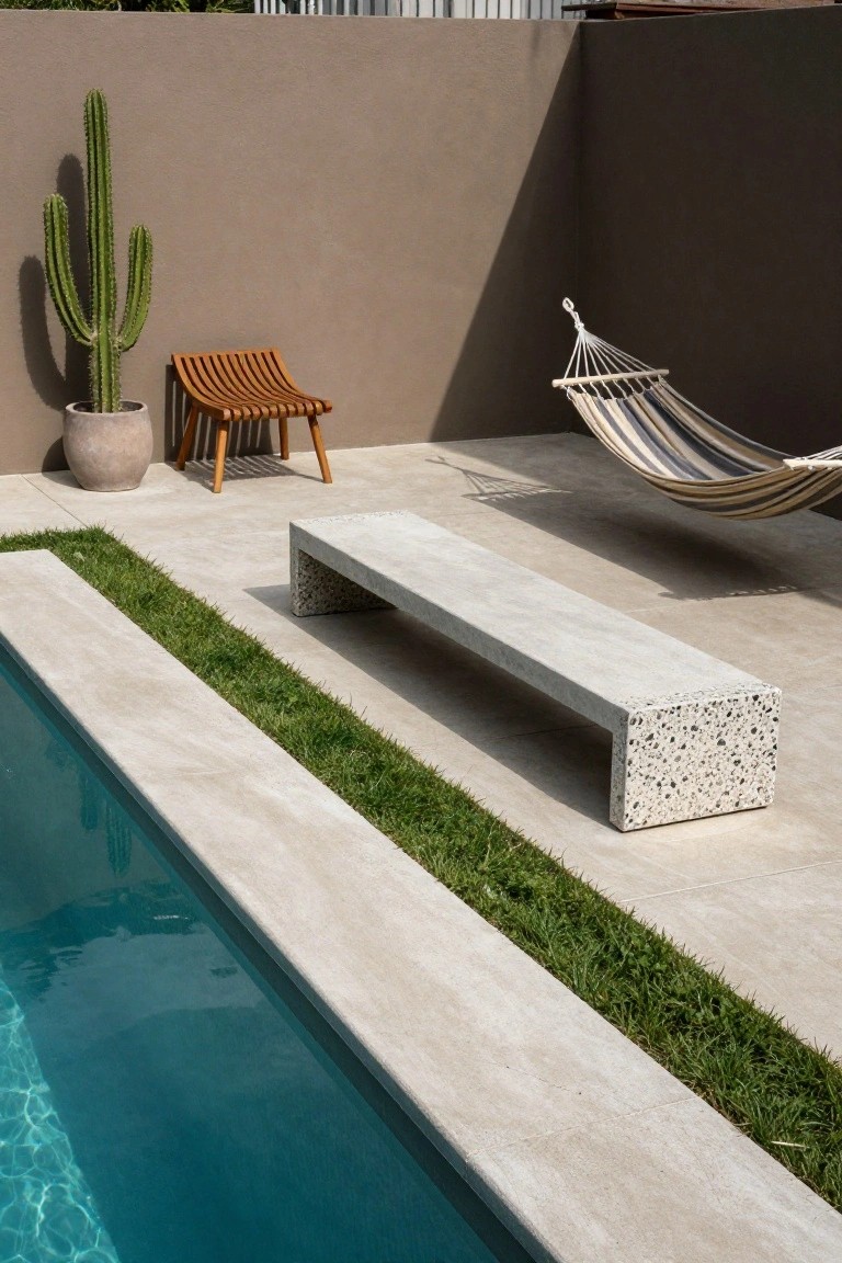 20 Durable Concrete Pool Landscaping Ideas That Feel Solid and Stylish Long gray concrete bench with terrazzo aggregate ends positioned parallel to an infinity-edge pool, separated by a narrow grass strip, with a hammock, wooden chair, and potted cactus on a travertine deck against a beige wall.