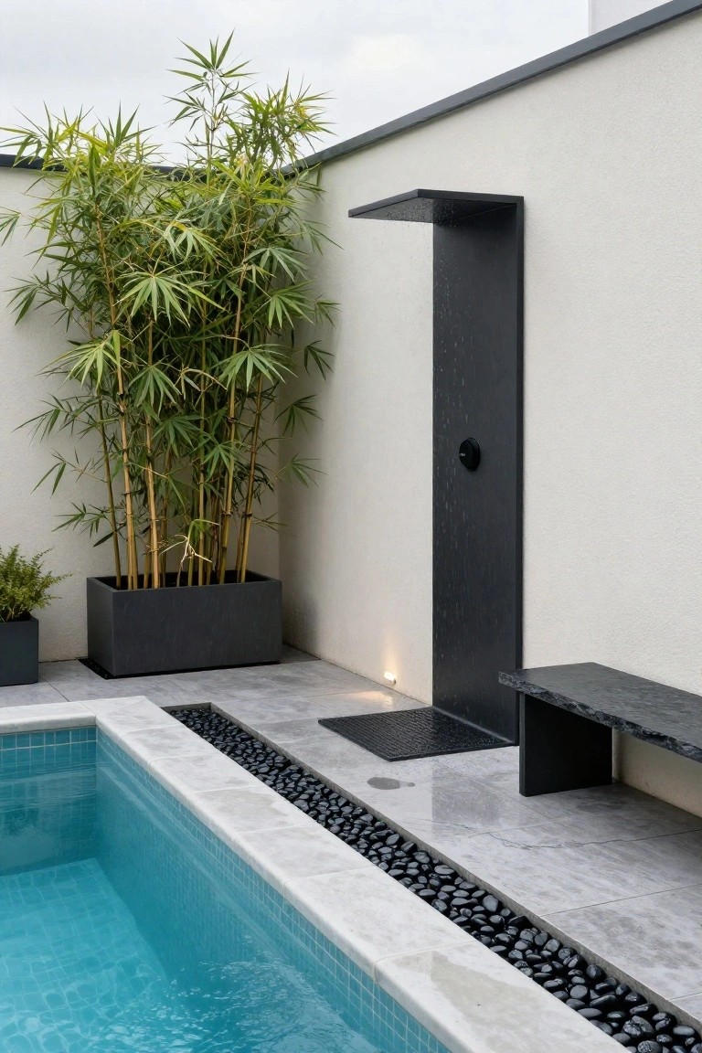 Modern black outdoor shower post with running water next to a turquoise mosaic-tiled pool edged in white stone and black pebbles, bamboo in a black rectangular planter for screening, stone bench, and gray tiled patio against a white wall.