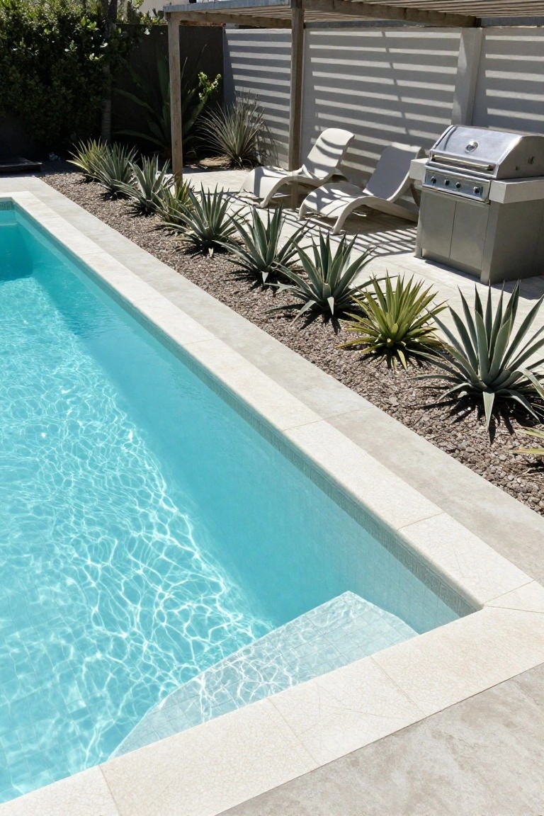 Turquoise pool with light beige waterline tiles seamlessly matching the coping, edged by agave plants and gravel, lounge chairs and stainless steel barbecue nearby under a pergola.
