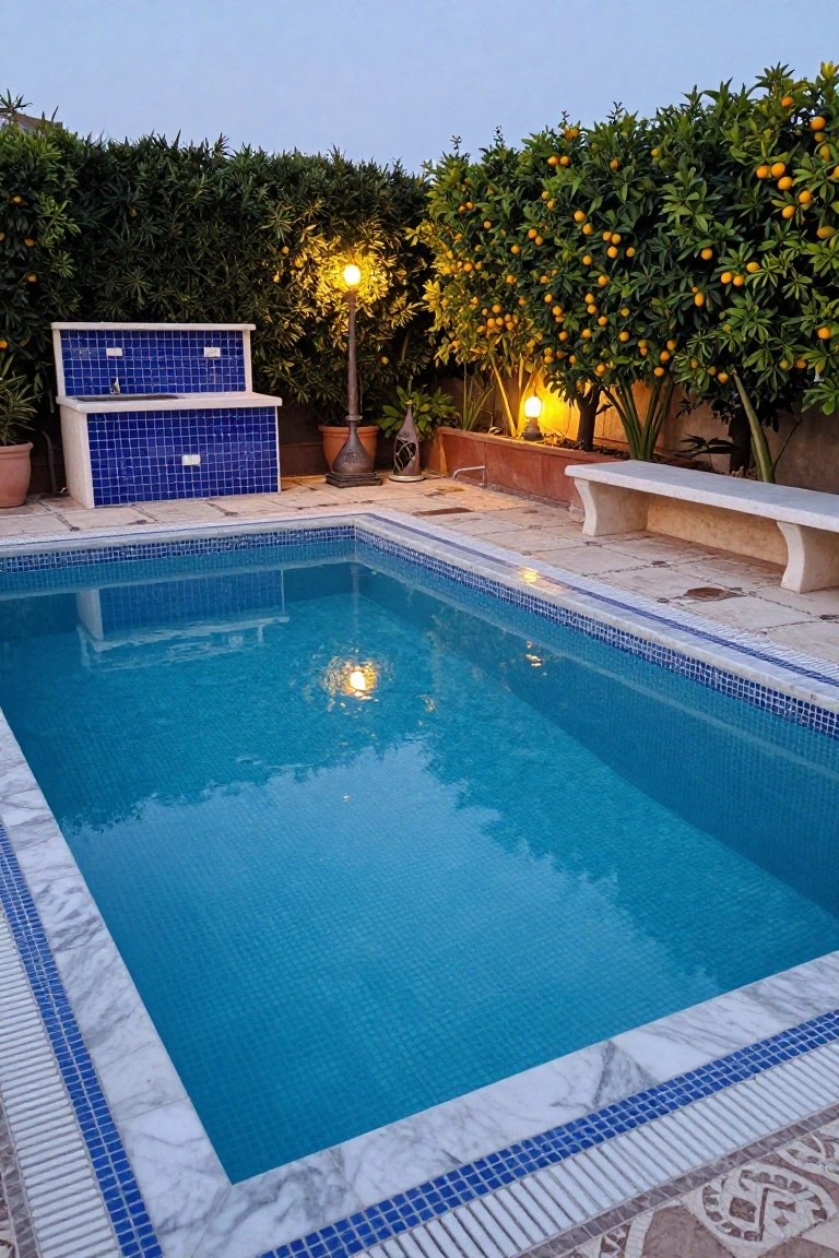 20 Eye-Catching Pool Edge Tile Ideas With A Defined Finish Rectangular swimming pool with blue mosaic tiles and white marble coping edged in blue mosaic, next to a stone bench, blue tiled sink unit, orange trees, hedges, and lamps in a walled backyard at dusk.