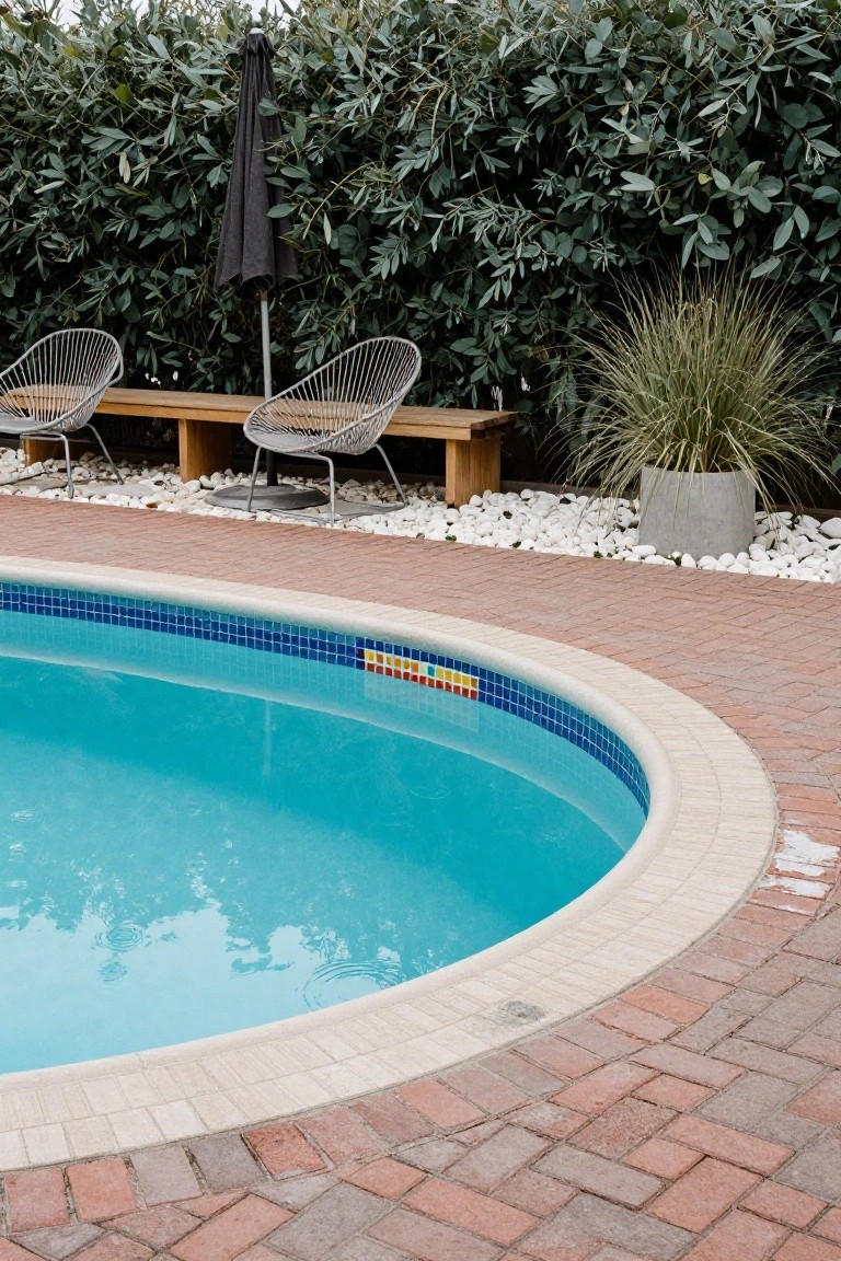 Curved blue-tiled swimming pool edged in white and brick pavers, with wooden bench and two wireframe chairs nearby, green hedges, ornamental grass in concrete pot, and white pebble ground cover.