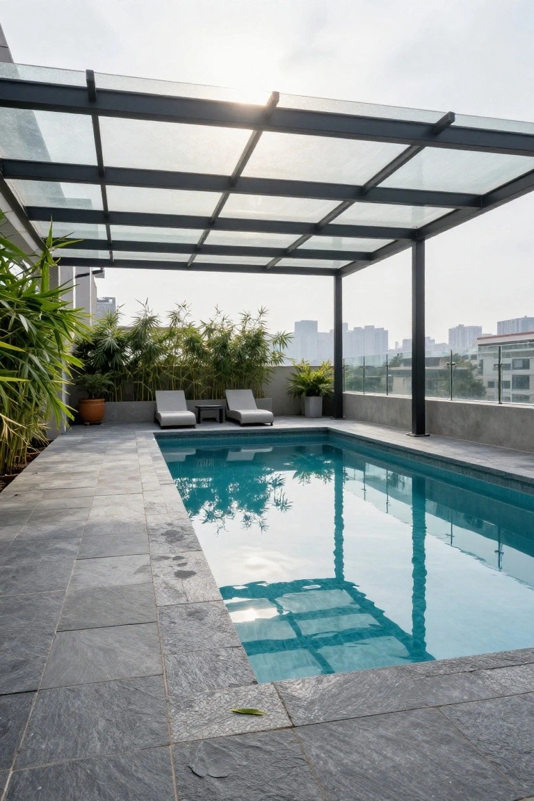 Rooftop pool surrounded by gray stone decking, with black-framed glass pergola overhead, two lounge chairs, potted plants, and distant city buildings.